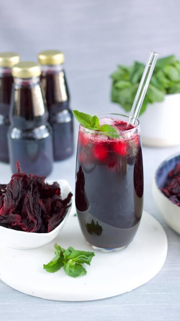 Jus de bissap rouge servi frais avec de la menthe, à côté de bouteilles et de fleurs d'hibiscus séchées